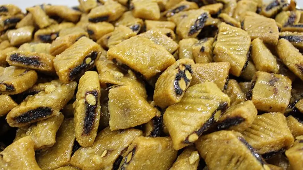 Magrood Libyan: Manisnya Warisan Kuliner dari Gurun Pasir 3 Dunia kuliner Timur Tengah sering kali identik dengan baklava atau kunafa yang sudah populer di lidah masyarakat global. Namun, jika kita menyelam lebih dalam ke pesisir Afrika Utara, terdapat satu permata tersembunyi yang menawarkan sensasi rasa berbeda bernama Magrood Libyan. Kudapan ini bukan sekadar kue kering biasa; ia adalah simbol keramah-tamahan masyarakat Libya yang telah diwariskan turun-temurun. Bayangkan sebuah kue berbasis semolina yang renyah di luar namun lumer di dalam, membawa rasa manis alami dari pasta kurma yang dipadukan dengan wangi air bunga mawar. Keunikan Magrood Libyan terletak pada keseimbangan tekstur dan aromanya yang mampu membawa siapa pun yang mencicipinya seolah sedang berada di tengah oasis yang tenang. Jejak Sejarah dan Filosofi di Balik Magrood Libyan Magrood, atau sering juga dieja sebagai Makroudh, memiliki akar sejarah yang kuat di kawasan Maghreb, mencakup Tunisia, Aljazair, hingga Libya. Di Libya, kue ini menempati kasta tertinggi dalam sajian hari raya seperti Idul Fitri atau acara pernikahan. Masyarakat setempat percaya bahwa menyajikan Magrood adalah cara menghormati tamu dengan memberikan yang terbaik dari hasil bumi mereka: gandum semolina dan kurma kualitas premium. Seorang pelancong fiktif bernama Omar pernah bercerita saat ia berkunjung ke sebuah rumah di Tripoli. Ia disambut dengan nampan berisi Magrood Libyan yang masih hangat dan aroma semerbak kayu manis. Baginya, setiap gigitan Magrood bukan hanya soal rasa manis, tapi tentang bagaimana sebuah bangsa mempertahankan identitasnya melalui resep yang tidak berubah selama berabad-abad. Tradisi ini tetap relevan bagi generasi milenial di sana yang mulai memodifikasi penyajiannya namun tetap mempertahankan bahan inti yang autentik. Kue ini mencerminkan adaptasi manusia terhadap alam gurun. Bahan-bahannya seperti kurma dan madu adalah sumber energi padat yang tahan lama di suhu panas. Oleh karena itu, Magrood Libyan sering kali menjadi bekal perjalanan bagi kafilah di masa lalu. Hingga saat ini, proses pembuatannya pun masih dianggap sebagai ritual sosial di mana para wanita berkumpul untuk membentuk adonan secara manual, menciptakan ikatan komunitas yang erat melalui dapur Blogger . Rahasia Bahan Utama yang Menciptakan Tekstur Sempurna Apa yang membuat Magrood Libyan berbeda dari kue kurma lainnya di dunia Arab? Jawabannya ada pada penggunaan tepung semolina kasar yang memberikan tekstur "berpasir" namun rapuh saat digigit. Berbeda dengan kue berbahan tepung terigu biasa, semolina memberikan karakter yang lebih kokoh dan mampu menyerap sirup madu dengan maksimal tanpa menjadi lembek. Selain semolina, elemen krusial lainnya adalah minyak zaitun atau mentega cair yang dicampurkan ke dalam adonan. Lemak ini berfungsi menciptakan lapisan yang kaya dan gurih. Berikut adalah beberapa komponen utama yang membangun keunikan rasa Magrood: Tepung Semolina: Memberikan tekstur unik yang renyah dan bergranula. Pasta Kurma: Sebagai isian utama yang memberikan rasa manis alami dan tekstur lembut. Air Bunga Mawar atau Oranye: Memberikan aroma floral yang menenangkan dan mewah. Madu dan Sirup Gula: Digunakan untuk merendam kue setelah dipanggang agar rasa manis meresap hingga ke dalam. Wijen: Sering ditaburkan di atasnya untuk memberikan aroma kacang yang sangrai. Proses pencampuran bahan ini membutuhkan insting yang tajam. Jika terlalu banyak air, adonan akan menjadi keras; jika terlalu sedikit, kue akan hancur saat dipotong. Inilah mengapa keahlian tangan seorang pembuat kue sangat menentukan hasil akhir dari Magrood yang sempurna. Seni Membentuk dan Memotong Magrood secara Tradisional Keunikan visual Magrood Libyan terletak pada bentuk belah ketupatnya yang presisi. Nama "Magrood" sendiri secara harfiah merujuk pada teknik memotong adonan secara diagonal. Proses ini dimulai dengan meratakan adonan semolina, lalu meletakkan gulungan pasta kurma di tengahnya sebelum digulung kembali hingga membentuk silinder panjang. Setelah gulungan terbentuk, pengrajin kue biasanya akan menghias bagian atas adonan dengan alat khusus yang disebut "manqash" untuk membuat pola ukiran. Pola ini bukan sekadar hiasan; celah-celah kecil hasil ukiran tersebut berfungsi untuk menangkap sirup madu agar meresap lebih baik. Setelah diukir, barulah adonan dipotong-potong menjadi bentuk berlian yang ikonik. Persiapan Adonan: Mencampur semolina dengan lemak hingga teksturnya seperti pasir basah. Pengolahan Isian: Menghaluskan kurma dan mencampurnya dengan sedikit kayu manis serta minyak agar lentur. Penyatuan: Memasukkan isian ke dalam adonan dan menggulungnya dengan rapi. Dekorasi: Mengukir permukaan adonan dengan motif tradisional. Pemotongan: Memotong adonan secara miring untuk mendapatkan bentuk belah ketupat. Langkah-langkah sistematis ini memastikan setiap keping Magrood Libyan memiliki proporsi isian dan kulit yang seimbang. Konsistensi dalam memotong sangat penting agar semua kue matang secara merata saat dipanggang di dalam oven bersuhu sedang. Teknik Pemanggangan dan Sentuhan Akhir yang Manis Berbeda dengan beberapa versi di negara tetangga yang digoreng, Magrood Libyan lebih sering dipanggang untuk mendapatkan hasil yang lebih ringan dan tidak terlalu berminyak. Proses pemanggangan dilakukan hingga kue berubah warna menjadi keemasan yang cantik. Namun, petualangan rasa Magrood Libyan belum selesai setelah keluar dari oven. Sesaat setelah matang, kue yang masih panas langsung dicelupkan ke dalam sirup madu atau "atsila" yang dingin. Perbedaan suhu ini menciptakan efek vakum yang menarik sirup masuk ke dalam pori-pori semolina. Hasilnya adalah kue yang terlihat berkilau di luar dan terasa sangat juicy di dalam. Teknik perendaman ini membutuhkan waktu yang pas. Jika terlalu sebentar, bagian tengah kue akan terasa kering. Jika terlalu lama, kue berisiko kehilangan tekstur renyahnya. Banyak pembuat kue profesional di Libya yang menambahkan perasan jeruk nipis ke dalam sirupnya untuk memberikan sedikit rasa segar guna menyeimbangkan rasa manis yang dominan dari kurma dan madu. Mengapa Magrood Relevan untuk Gaya Hidup Modern Bagi generasi muda, Magrood Libyan mulai dipandang sebagai alternatif camilan sehat dibandingkan biskuit pabrikan yang penuh pengawet. Kandungan kurma yang kaya akan serat dan energi alami menjadikan kue ini teman yang sempurna untuk secangkir kopi hitam tanpa gula atau teh hijau di sore hari. Inovasi juga mulai bermunculan di kafe-kafe modern di Tripoli maupun di kalangan komunitas diaspora Libya di luar negeri. Ada yang mulai menambahkan kacang almond atau pistachio ke dalam isian kurma untuk menambah tekstur renyah yang lebih modern. Namun, bagi para purist, keaslian Magrood yang menggunakan 100% kurma murni tetap tidak terkalahkan. Selain itu, kemasan Magrood yang kini lebih estetik menjadikannya pilihan hadiah atau hantaran yang elegan. Di tengah tren "back to basics", kuliner tradisional yang memiliki cerita kuat seperti Magrood mendapatkan tempat spesial di hati konsumen yang mencari otentisitas dalam setiap makanan yang mereka konsumsi. Menikmati Magrood sebagai Pengalaman Budaya Menikmati Magrood Libyan adalah sebuah pengalaman sensorik yang lengkap. Dimulai dari melihat bentuknya yang geometris, mencium aroma bunganya, hingga merasakan tekstur semolina yang unik di lidah. Ini adalah cara termudah bagi kita untuk mengenal kebudayaan Libya tanpa harus melintasi samudera. Setiap gigitan kue ini mengandung sejarah panjang peradaban Mediterania dan Afrika Utara. Magrood bukan hanya tentang rasa manis, melainkan tentang ketekunan dalam proses dan rasa bangga akan warisan leluhur. Dengan segala keunikan kuliner Magrood Libyan yang telah dibahas, tidak mengherankan jika kudapan ini tetap bertahan dan terus dicintai melintasi zaman dan batas negara. Sebagai penutup, eksplorasi kuliner dunia memang tidak akan pernah ada habisnya selama kita mau membuka diri pada rasa-rasa baru yang autentik. Magrood adalah pengingat bahwa kebahagiaan sering kali datang dalam bentuk yang sederhana, seperti sepotong kue kurma yang dibuat dengan cinta dan tradisi yang terjaga. Baca fakta seputar : Culinery Baca juga artikel menarik tentang : Es Krim Turki: Sensasi Kenyal Unik yang Bikin Penasaran Dunia